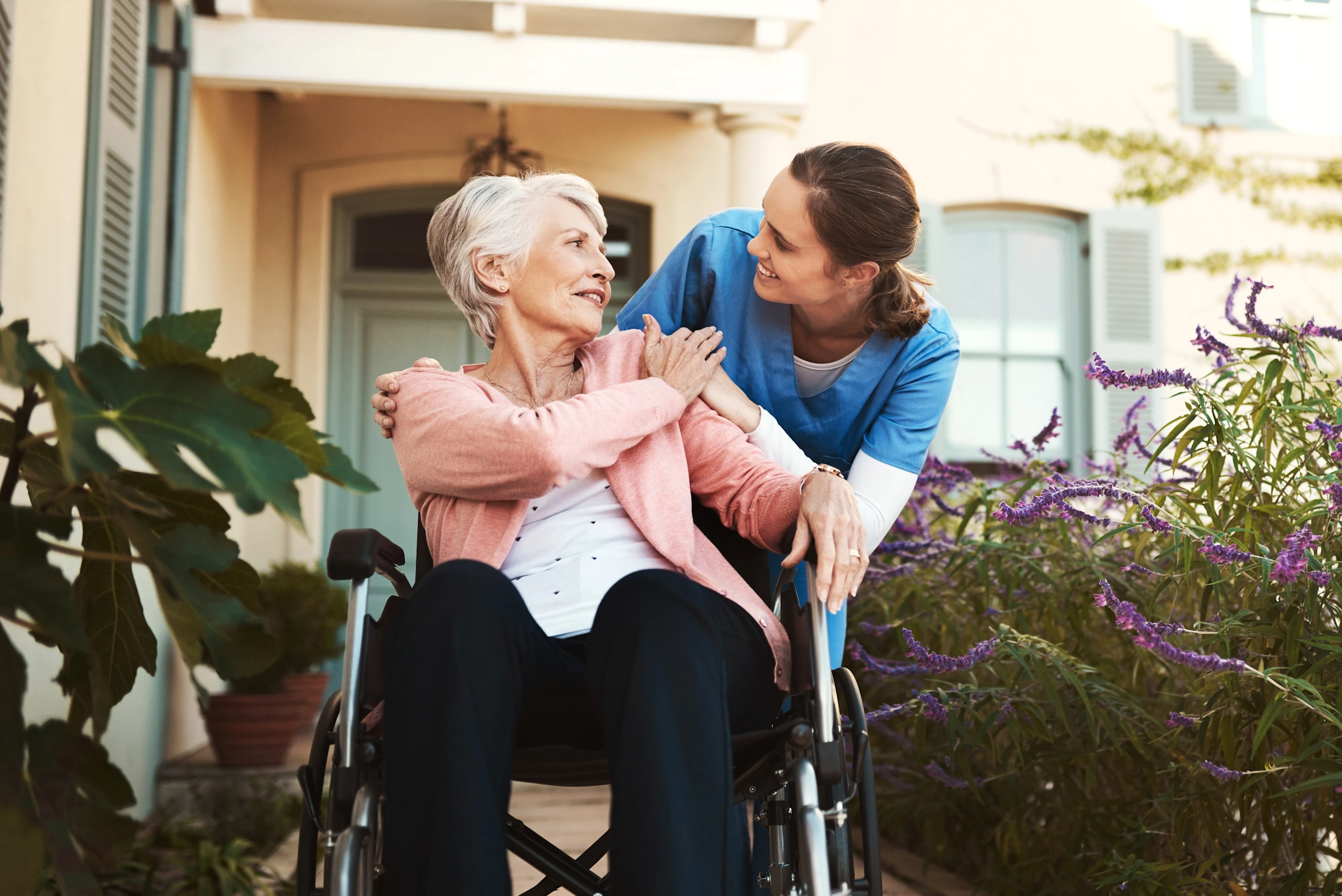 A compassionate caregiver supports a smiling senior woman in a wheelchair outdoors.